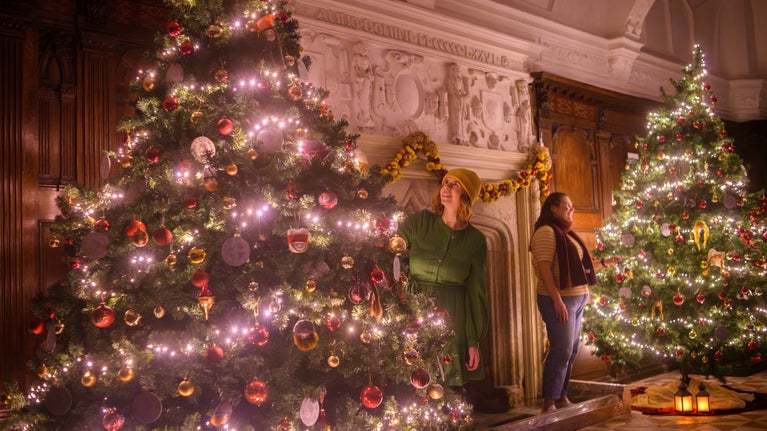 Two large Christmas trees decorated with red and gold baubles and fairy lights stand either side of a large fireplace. Two women are dressed in winter clothes admiring the trees. There is wood panelling and plasterwork in the background and lanterns on the ground.